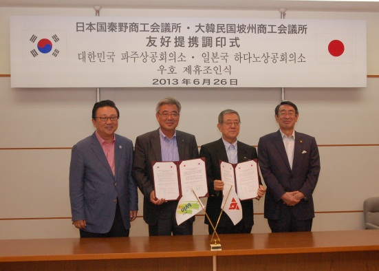 「友好提携 調印式」と書かれた横断幕の下の室内で、スーツ姿の4人の男性がテーブルの前に立ち、中央の2人がそれぞれ両手で調印された協定書を持ち記念撮影している写真