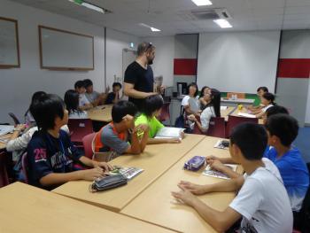 教室の室内で、机を向かい合わせにして座る小学生の間に、外国人教師が立ち、小学生と話している写真