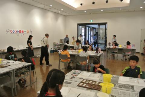 広い部屋で、新聞紙を敷いた複数のテーブルに大人と子供たちが椅子に座り、制作活動を行っている様子の写真