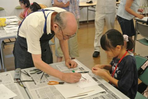 エプロンを着用した男性が、新聞紙を敷いた机の席に座っている女の子に対し、道具を使いながら作業の手伝いを行っている様子の写真