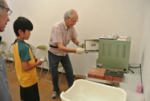 男性が白い手袋を着用し、緑色の小型電気炉の扉を開けて作業している様子を、少年が見守っている写真