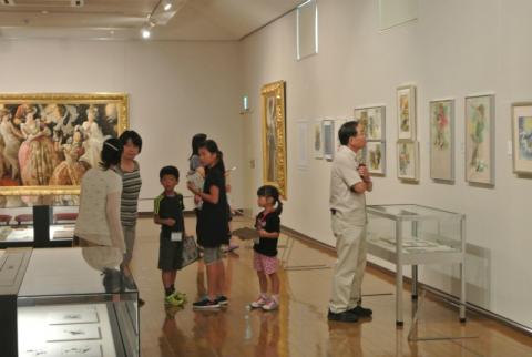 明るい照明と木目の床の美術館の展示室で、壁にかけられた大きな絵画や複数の額装された作品を、家族連れなどの複数の来館者が鑑賞している写真