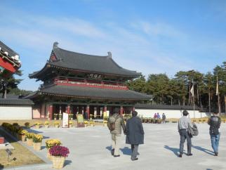 青空の下、黒い瓦屋根が特徴的な韓国の伝統的な建築物である烏竹軒と書かれた門を、数名の観光客らしき人々が広場を横切って歩いていく様子の写真