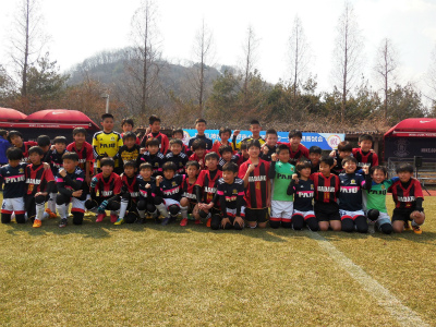 芝生のグラウンドで、赤や黒色などのユニフォームを着用した大勢の少年サッカーチームの選手たちが、笑顔で集合している写真