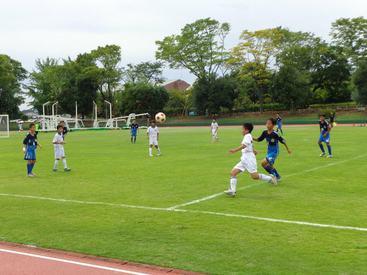 緑の芝生のサッカー場で、白いユニフォームと紺色のユニフォームを着た複数の少年たちが、試合を行っている様子の写真