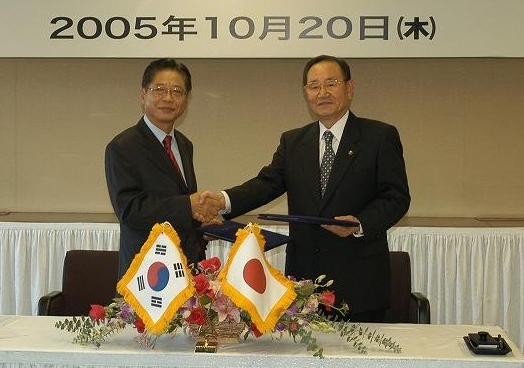 日韓の国旗をあしらった装花が置かれたテーブルの前で、濃色のスーツを着た2人の男性が書類を挟んで笑顔で握手しており、背景には「2005年10月20日（木曜日）」の日付が示された調印式の写真