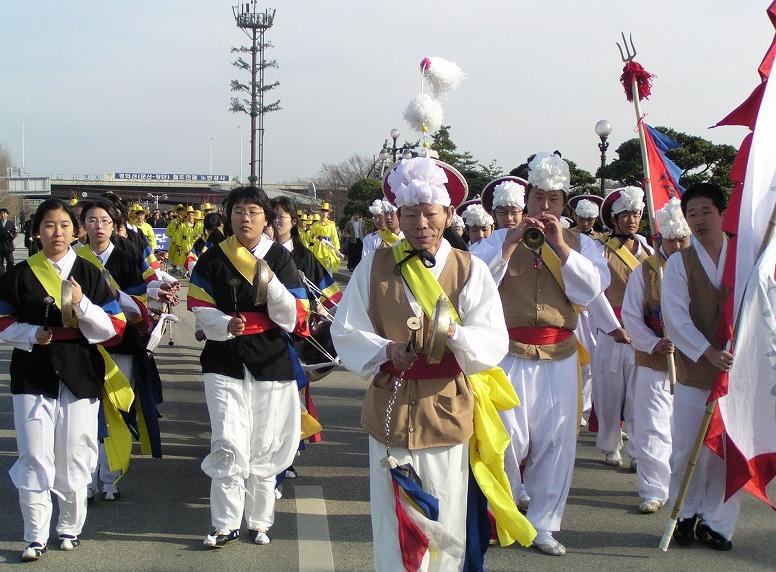 韓国の伝統的な農楽の衣装を身にまとった人々が、鉦やラッパなどの楽器を演奏しながら屋外の道路を整然と練り歩いており、白い花飾りの帽子や鮮やかなたすきが印象的なパレードの写真