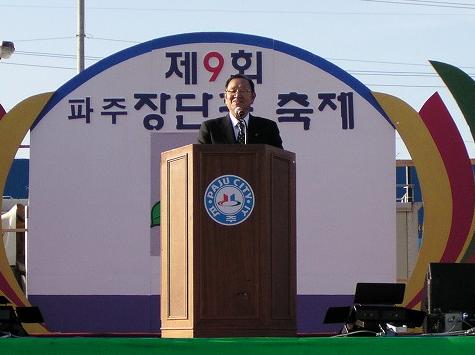 PAJU CITYのロゴが入った演台に立ち、ハングルで第9回と書かれたアーチ状の看板を背にして、スーツ姿の男性がマイクに向かいスピーチをしている屋外イベントの写真