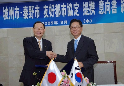 日韓の国旗が置かれたテーブルの前で、スーツ姿の2人の男性が笑顔で握手を交わしており、背後の青い横断幕には「坡州市・秦野市 友好都市協定 提携意向書」書かれた調印式の写真