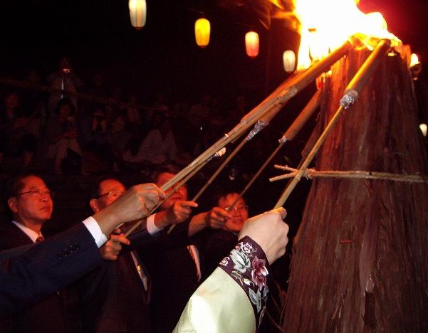 暗闇の中、伝統的な衣装やスーツ姿の数人が、火のついた松明や長い竹竿を高く積み上げられた護摩壇に向かってかざし、炎を点火している様子の写真