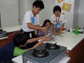 調理室で、4人の学生がガスコンロの前に置かれたテーブルの上で、ボウルに入った食材を混ぜている写真