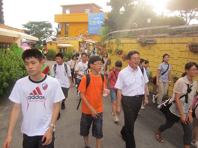 黄色の建物や壁がある街中で引率者の男性と、リュックを背負った複数の学生たちが歩いている様子の写真
