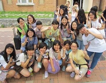 赤茶色のレンガ造りの建物前で、大勢の女子学生たちがピースをし記念撮影している写真