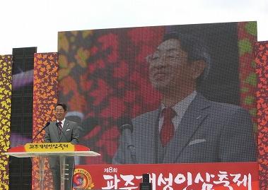 背景の大型ビジョンに、スーツを着用しマイク付きの演台の前に立って挨拶をしている男性のアップが映し出されている屋外の特設ステージの写真