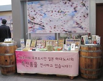 屋内の展示ブースで、桜の写真が背景に飾られ、2個のたるの間に設置されたテーブルには様々な特産品が並べられ、テーブル前面の垂れ幕には韓国語で書かれた横断幕があるブースの写真