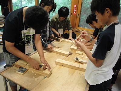 作業台の上で、学生たちが鉋や木片を使ってはし＆はし袋作りをしている様子の写真