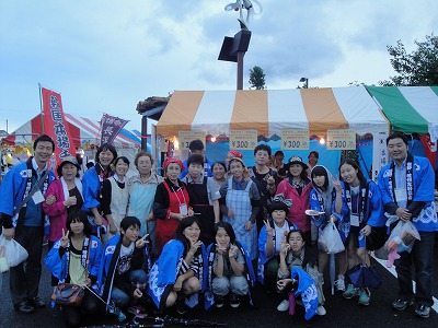 法被を着た人やエプロン姿の地元の人々や学生たちがオレンジと白色のストライプ柄のテントの前で記念撮影している写真