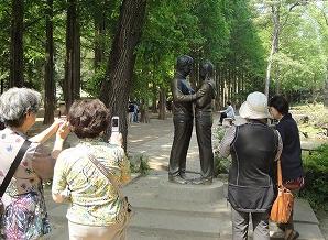 韓国の並木道の公園内にある、抱き合う男女の銅像を、数名の年配の女性の観光客がスマートフォンやカメラで熱心に撮影している様子の写真