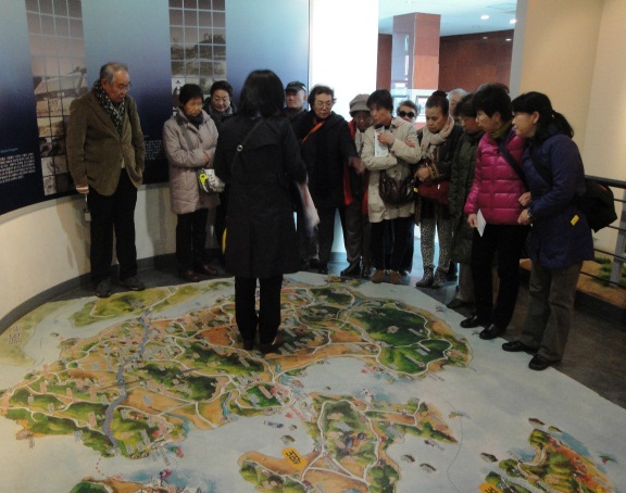 屋内の展示スペースで、数名の訪問団員と思われる人々が、床に大きく描かれた地図や模型のようなものを取り囲み、案内役と思われる女性の説明を熱心に聞いている様子の写真