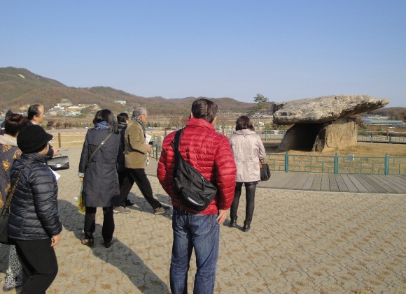 青空と低い山々を背景に、訪問団員と思われる数名の男女が、巨大な石の蓋と支石からなる特徴的な形状のコインドル（支石墓）を屋外の史跡で見学している様子の写真