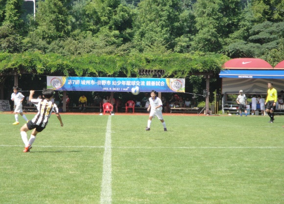 背景の藤棚に「第7回 城州・茅野市 幼少年サッカー交流親善試合」と書かれた横断幕が掲げられている緑の芝生のサッカー場で、ユニフォームを着た数人の少年たちが試合をしている写真