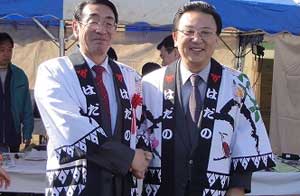 スーツの上に「はだの」の文字や様々な柄が描かれた白黒の法被を着用した二人の男性が笑顔で記念撮影している写真