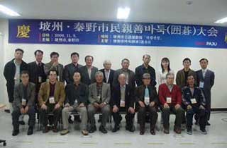 「慶 坡州・秦野市民親善バドク(囲碁)大会」と書かれた横断幕の前で、スーツやカジュアルな服装の男性を中心に、総勢19名の両市の親善訪問団員が集まり、記念撮影している様子の写真
