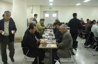 屋内の広い会場で、多くの年配の男性たちが向かい合って座り、バドク（囲碁）の対局を熱心に行い、周囲では他の参加者たちがその様子を見ている交流会の様子の写真