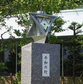 縦書きで「平和の灯」と書かれた白いプレートが貼られたグレーの御影石でできた台座の上に、ステンレス製の三角形が組み合わされた平和の灯モニュメント設置されている写真