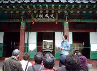 「文成祠」と書かれた額が掲げられた朝鮮半島の伝統的な建築物である祠の前で、男性のガイドと思われる人物が、数名の年配の観光客らしき人々に説明をしている様子の写真