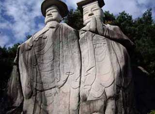 青空を背景に、山や森の中にある、二体の巨大な石仏が並び立つ姿を、やや下から見上げるようにして捉えた迫力のある写真