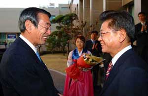 スーツ姿の二人の男性が笑顔で向かい合って握手をしており、その間にチマチョゴリを着た女性が花束を持って立っている歓迎の場面の写真