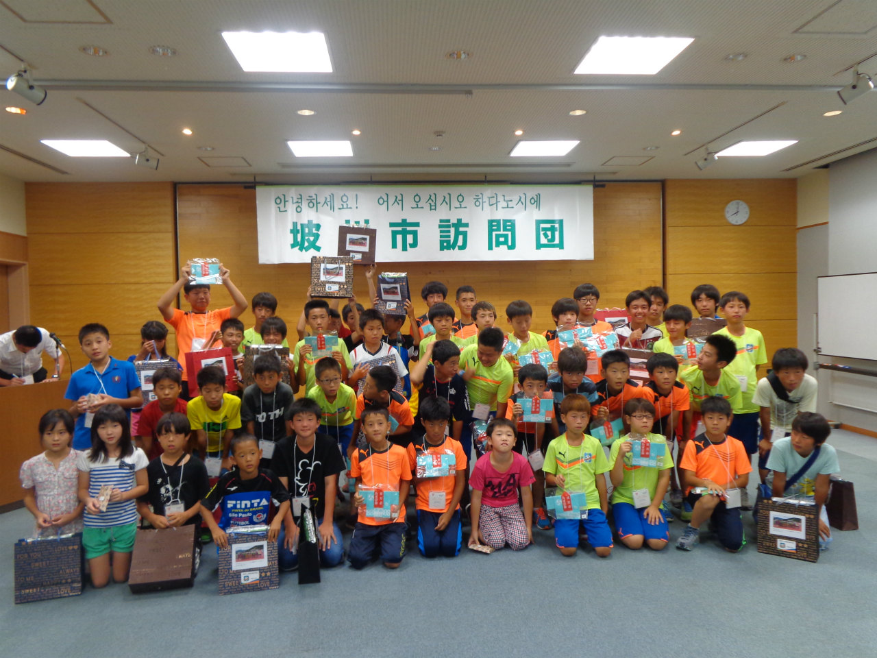 韓国語と日本語で「坂州市訪問団」と書かれた横断幕の下でオレンジや黄緑のサッカーユニフォーム姿の少年たちが記念撮影している写真