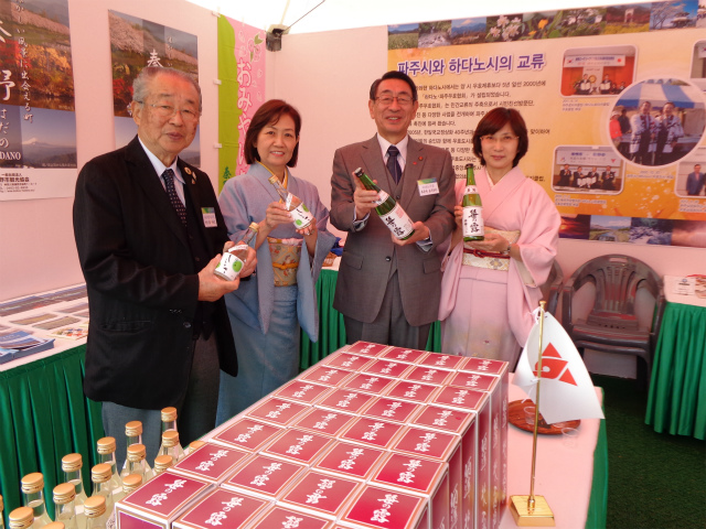 男性2名がスーツ、女性2名が和服を着用し、それぞれお酒の瓶を手に並んで立っており、手前のテーブルには多数の箱や瓶が展示され、背景には秦野市と坡州市の交流を紹介するポスターが貼られた特産品ブースの写真