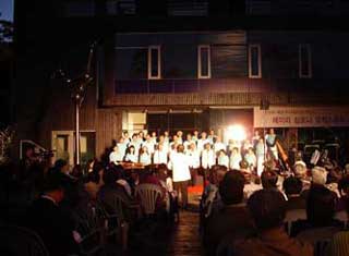 夜間の屋外特設ステージで、白い衣装を着用した数十名の合唱団が歌を披露し、多くの観客が椅子に座ってその演奏を鑑賞している様子の写真