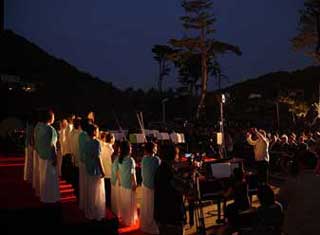 屋外の特設ステージで、後列に白いロングスカートと水色のジャケットを着用した数十名の女性、前列に白のジャケットを着用した数十名の男性の混成合唱団が、夜間の暗闇の中でスポットライトを浴びながら歌声を披露している様子の写真