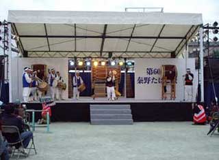 「第60回 秦野たばこ祭り」と思われる文字が書かれた屋外の特設ステージ上で、数名の演者が白い衣装と青いベストを着用し、韓国の太鼓や笛の演奏を披露している様子の写真