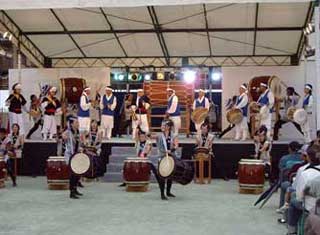 白い屋根の特設ステージで、白い股引と青いベストを着用した数十名の韓国の演者たちと、ステージ下の秦野観光和太鼓の演者たちと競演して演奏を披露している写真