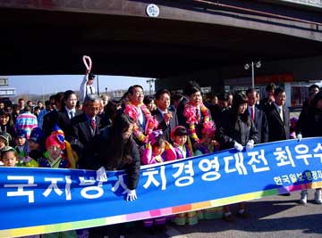 高架下で、民族衣装を着た子供たちを含む多くの群衆と、ピンクのレイをかけられた複数のスーツ姿の男性が、青い横断幕を持ってパレードに参加している様子の写真