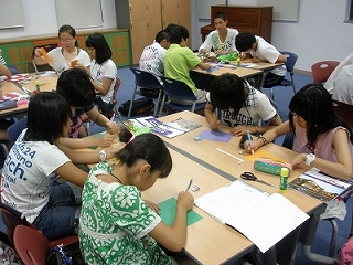 学生たちが2人掛けの机を囲み向い合わせにして座り、ハサミやのり、色鉛筆などが置かれた机で色画用紙に書いている様子の写真