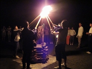 複数の若者が中央に組まれた焚き火の周りを囲み、長い棒を使って火を点けようとしている点火式の様子の写真