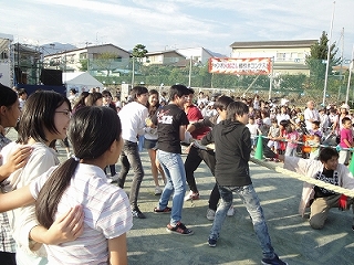 屋外でたくさんの観客や参加者が見守る中、学生たちがロープを力いっぱい引っ張り合う綱引き競技をしている様子の写真