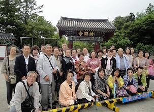 「佛國寺」と書かれた額が中央に掲げられた瓦屋根の門を背景に、多数の男女の訪問団員たちが前に並べられた黒と黄色の柵に囲まれて記念撮影をしている集合写真