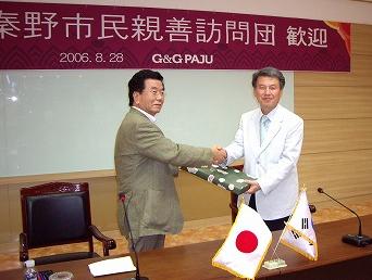 2006年8月28日、「秦野市民親善訪問団 歓迎 G&G PAJU」と書かれた横断幕の下、秦野市の団長と思われる男性と、パジュ市の関係者と思われる男性が、握手を交わしながら贈呈品を受け渡している様子の写真