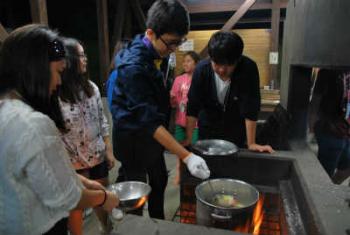 野外の炊事場で、数人の若者たちがかまどの上に置かれた大きな鍋をのぞき込んでおり、一人の男の子が鍋をかき混ぜている写真