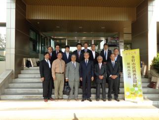 「ようこそ！秦野市民親善訪問団」と書かれた黄色の縦長の看板の横で、スーツ姿の男性を中心に、総勢14名の団員と思われる男性たちが、建物の階段の前で集合している様子の写真