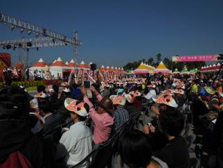 青空の下、多数の来場者がオレンジと白のテントが並ぶ賑やかなフェスティバル会場に集まり、ステージ上のイベントを個性的な帽子をかぶって見ている様子の写真