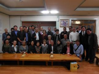 木製の低いテーブルが並んだ屋内の広間で、男性と女性の団員と思われる人々が、後列に立って前列に座る形で集合し、記念撮影をしている様子の写真