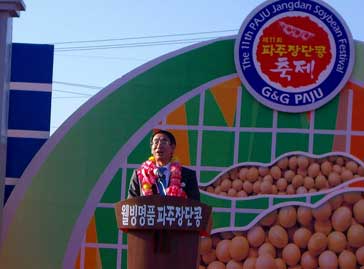 屋外のイベント会場のステージで、ピンクと白のレイをかけたスーツ姿の古谷市長がジャンダン豆が書かれたアーチ型の看板を背景に、マイクスタンドの前で挨拶をしている様子の写真