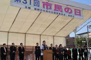 屋外のテント下特設ステージで、「第31回 市民の日」と書かれた横断幕の下、白い法被を着用した男性が演台で挨拶をしており、周囲にはスーツ姿の複数の関係者が並んでいる式典の写真